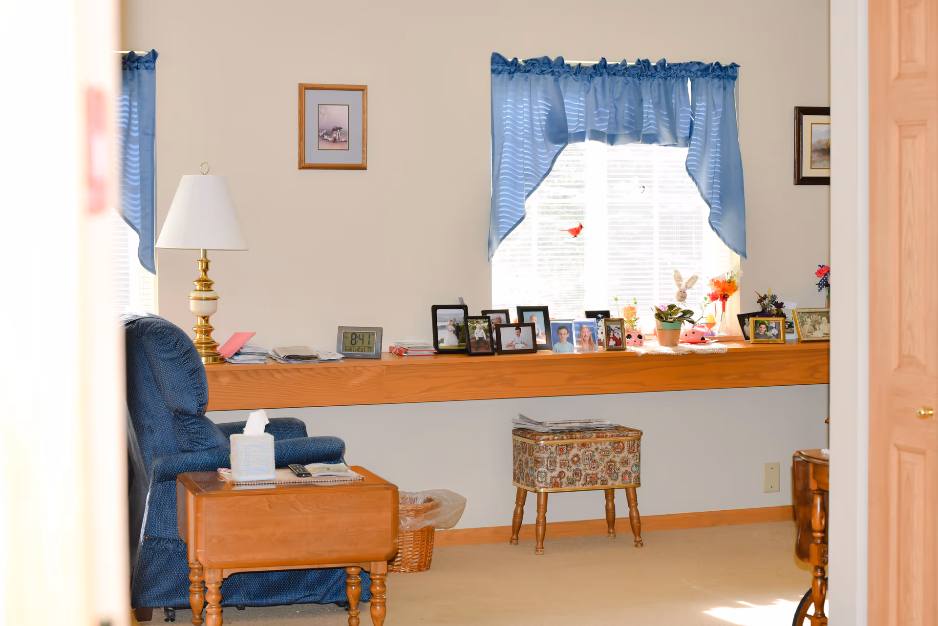 A cozy living room area with a blue upholstered recliner chair next to a wooden side table holding a tissue box and remote control. Behind the chair is a long wooden shelf beneath a window with blue curtains, displaying numerous framed photographs, small plants, and decorative items. A brass and white table lamp is also on the shelf. The room has beige walls and carpet, with a small patterned footstool under the shelf and a wicker wastebasket nearby.