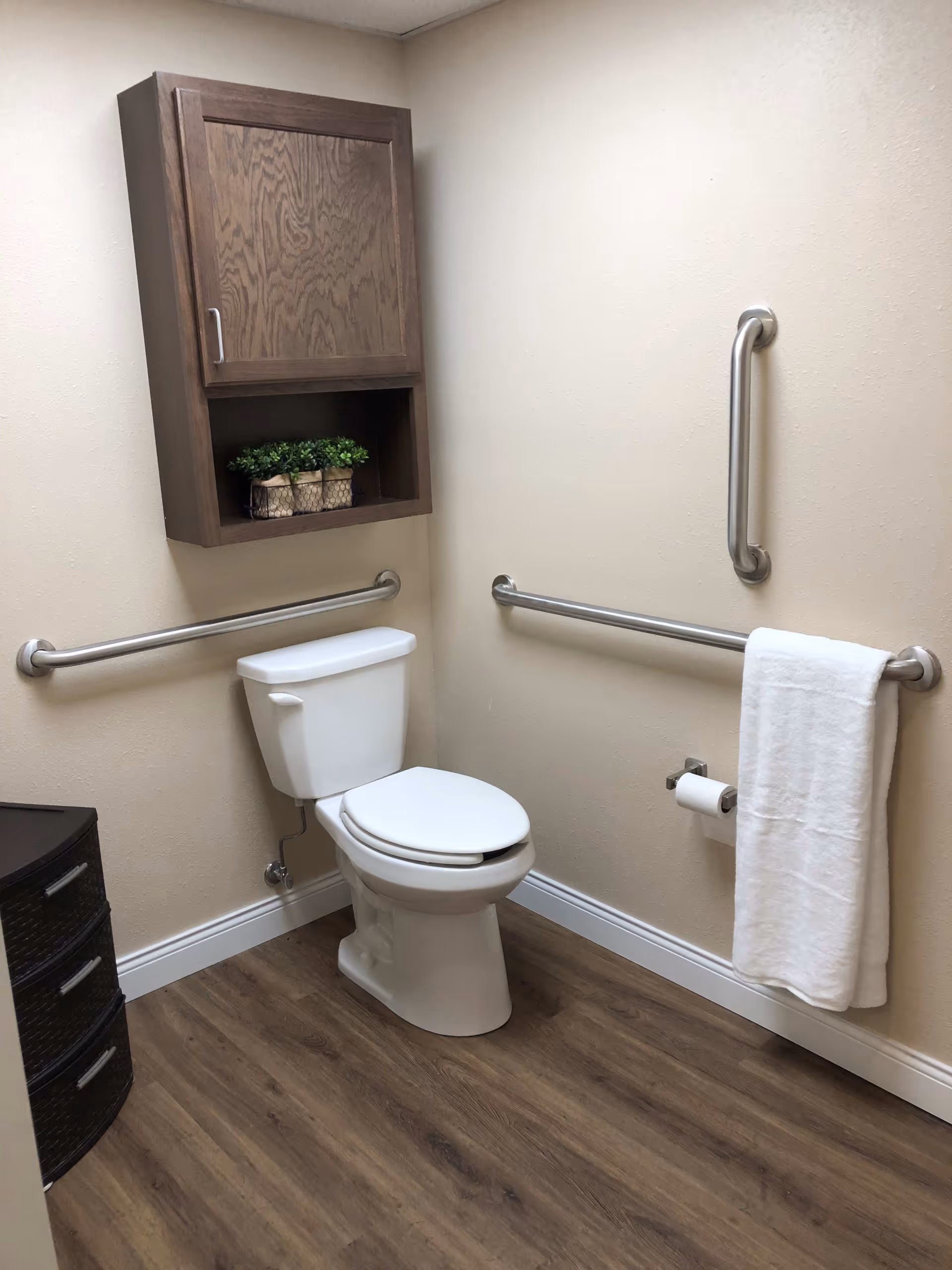 A clean and accessible bathroom with a white toilet, wooden cabinet mounted on the wall above the toilet, and stainless steel grab bars on the walls. A white towel hangs on one of the grab bars, and there is a small set of dark drawers in the corner. The floor is wood-patterned, and the walls are light beige.