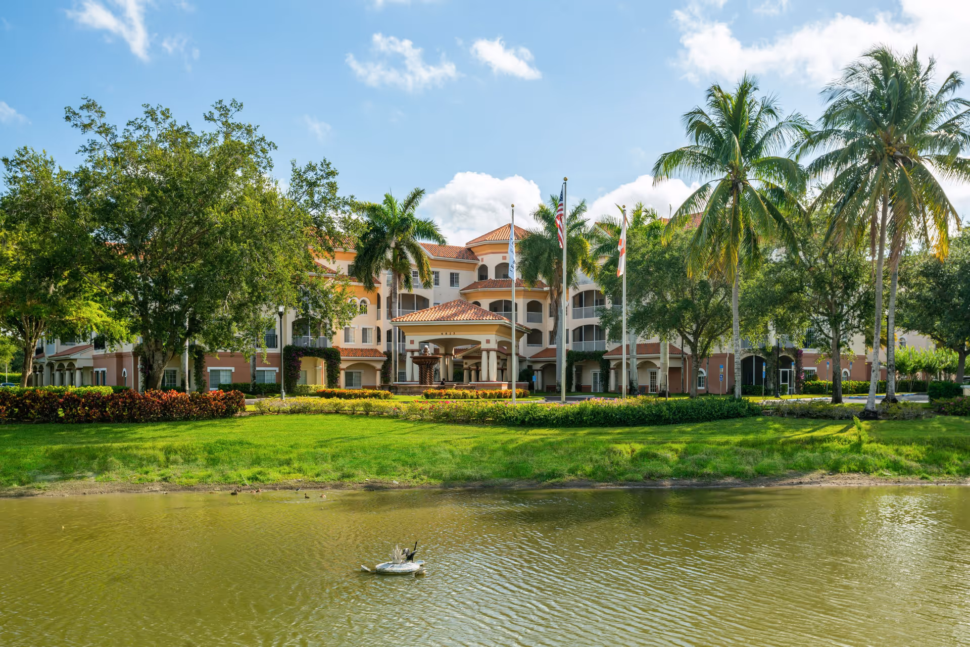 View of a senior living facility building with a terracotta roof, surrounded by palm trees and other greenery, with a pond in the foreground under a partly cloudy blue sky.