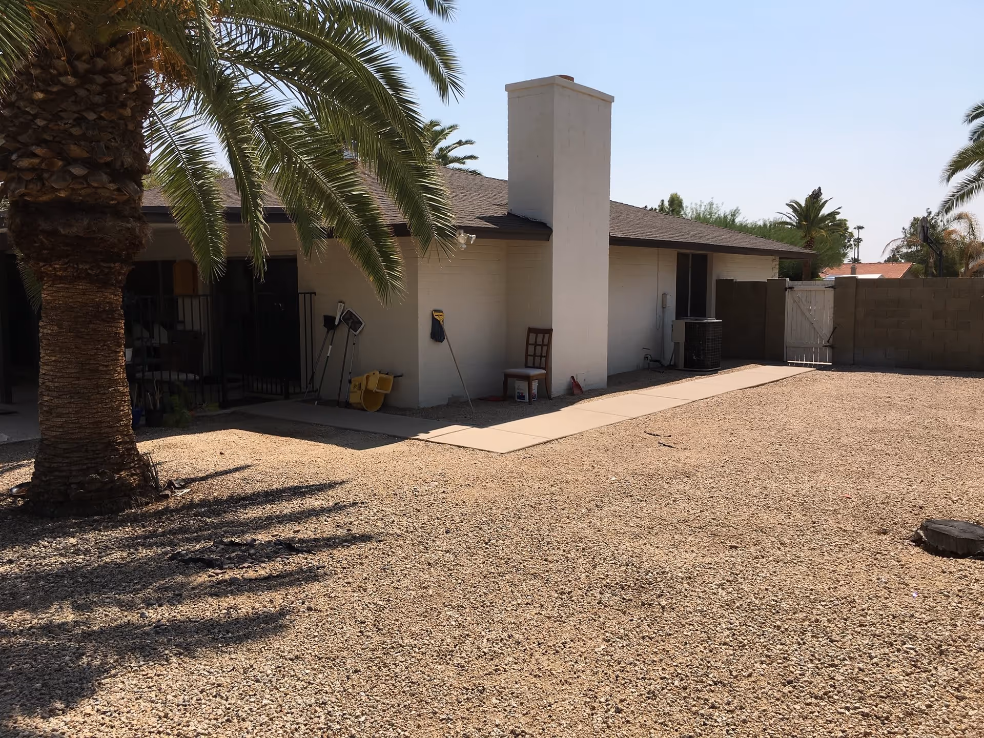 Outdoor view of a single-story building with a chimney, surrounded by a gravel yard and palm trees. There is a concrete walkway leading to a gate in a brick wall, and some cleaning tools and a chair are placed against the building wall.