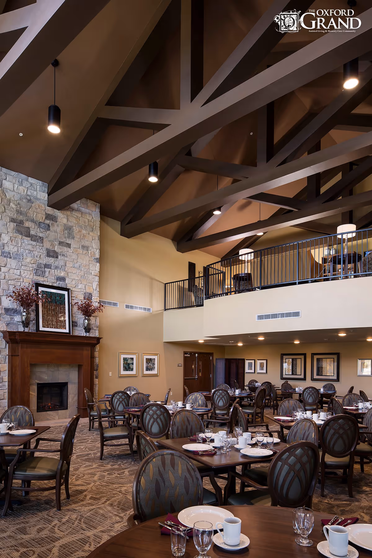 A spacious dining room with multiple round tables set with white plates, cups, glasses, and silverware. The room features a high vaulted ceiling with exposed dark wooden beams, a stone fireplace with a wooden mantle, and framed artwork on the walls. There is a mezzanine level with additional seating visible above.
