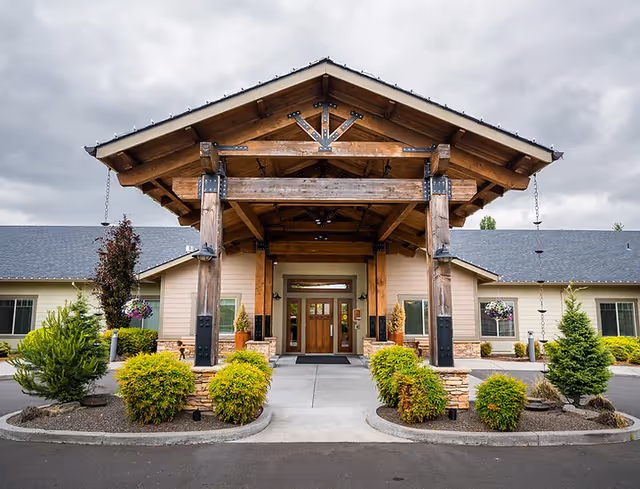Front entrance of Sunnyside Meadows Memory Care facility featuring a large wooden covered porch with exposed beams, surrounded by neatly trimmed bushes and small trees under a cloudy sky.