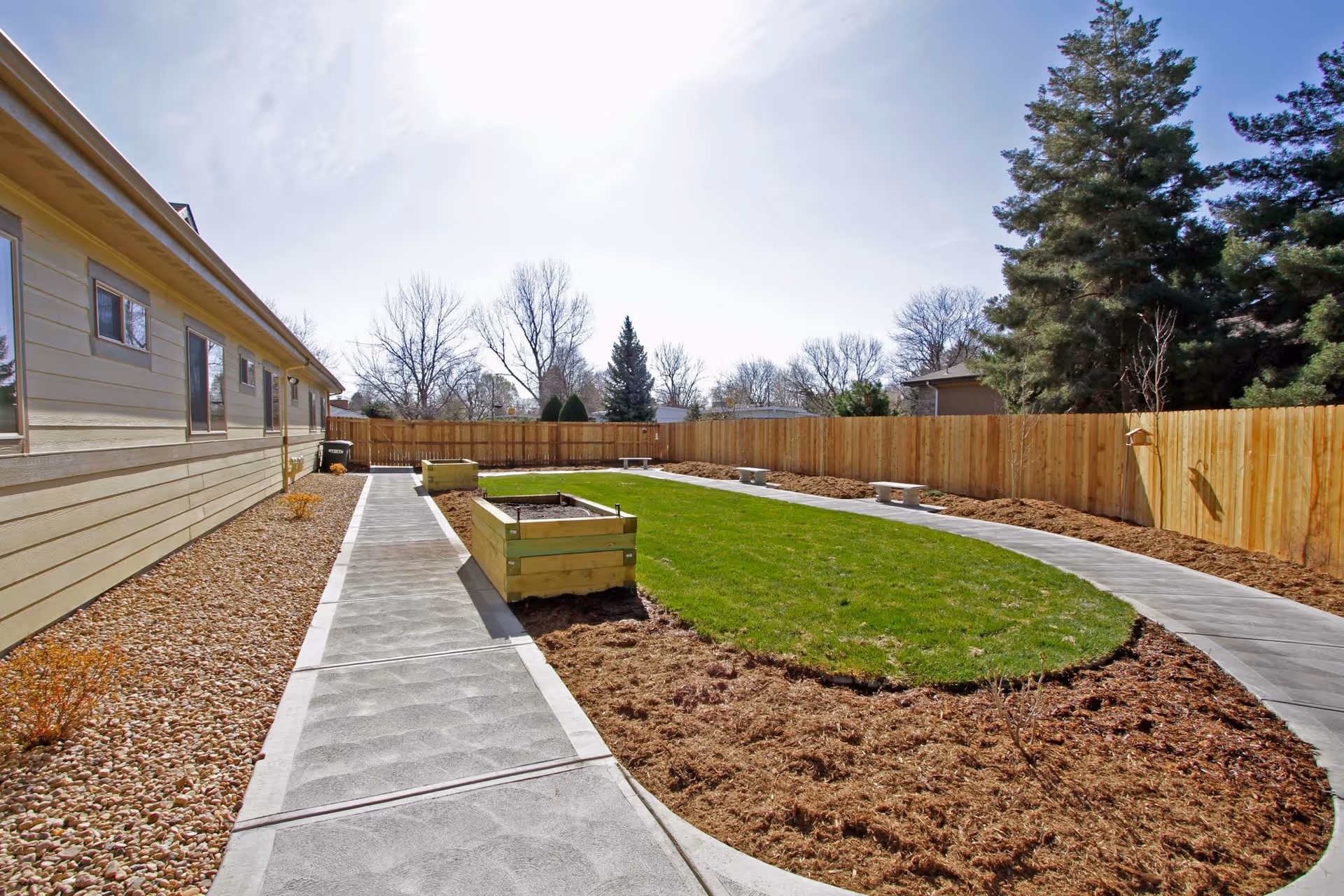 Outdoor area of a senior living facility with a concrete walkway surrounding a grassy oval lawn, wooden raised garden beds, and a wooden fence. Trees and shrubs are visible in the background under a clear sky.