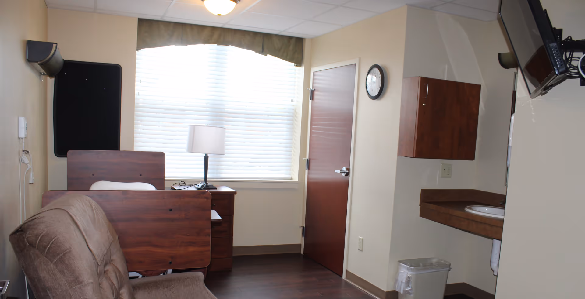 Small private resident room with a bed, armchair, window with blinds, bedside table and lamp, wall-mounted TV, and a sink area.