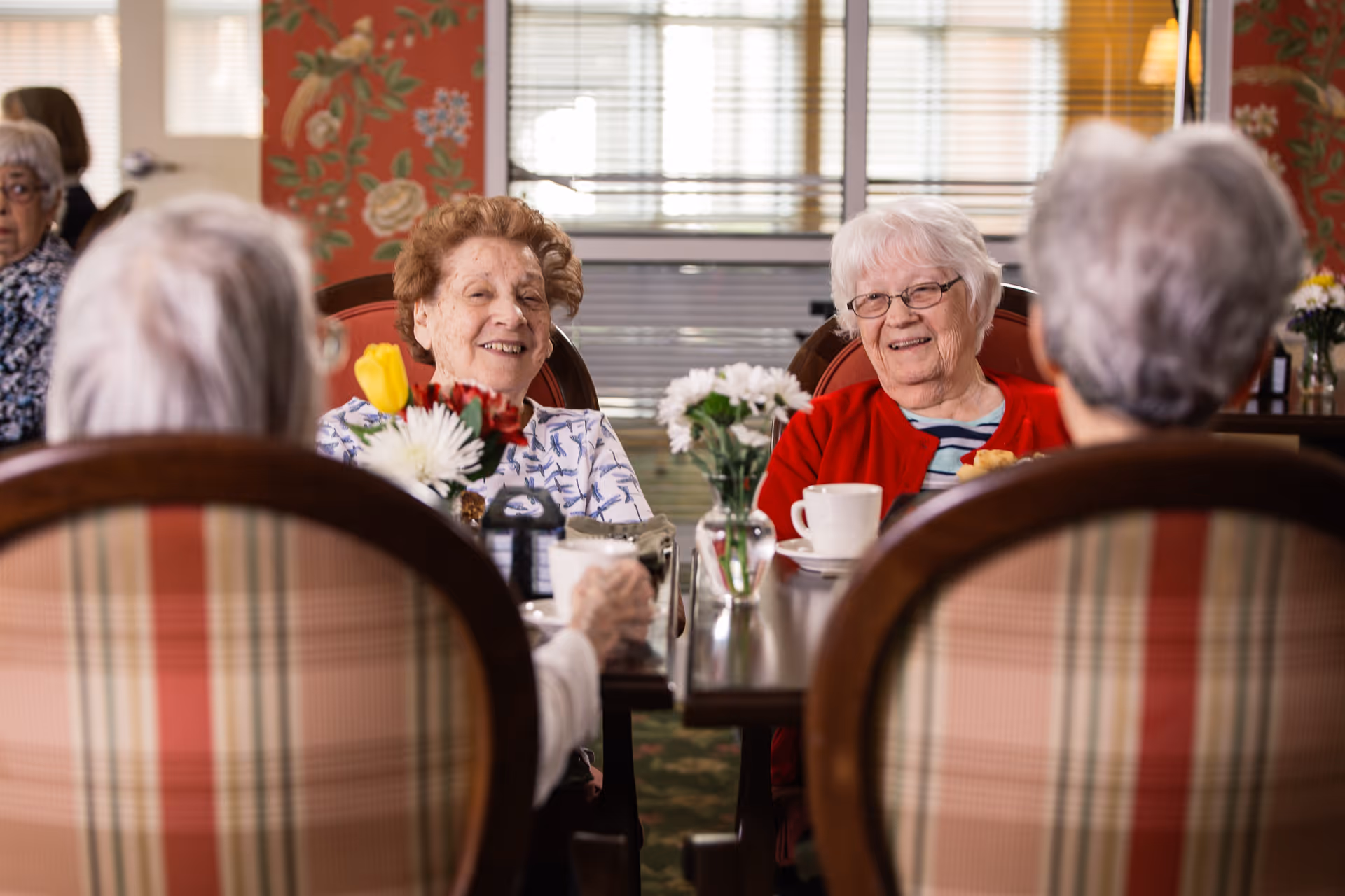 Several elderly women sit around a dining table with flowers and teacups, smiling and chatting in a communal dining room.