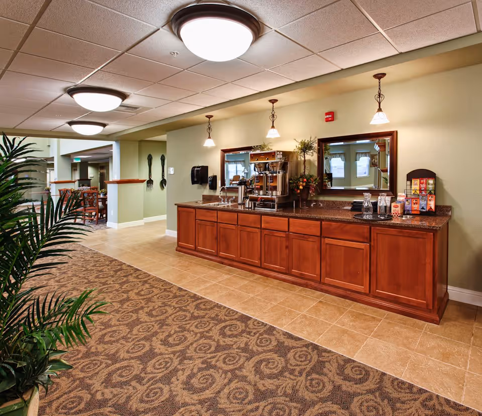 Interior view of a senior living community area featuring a long wooden counter with a coffee machine, cups, and a tea selection. The space has beige tiled flooring and patterned carpet, with green walls and ceiling lights. There is a large mirror above the counter and some decorative plants nearby. In the background, there are dining tables and chairs visible.