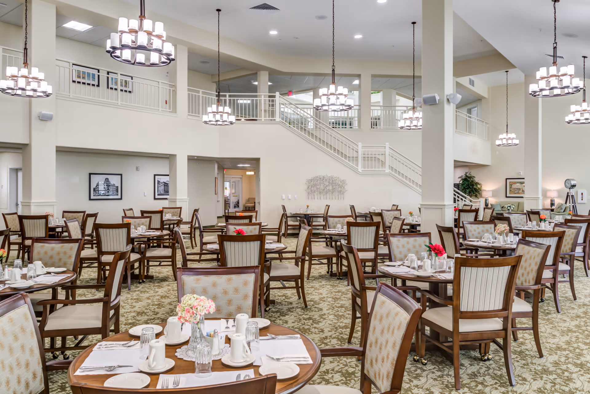 Spacious dining room with multiple round wooden tables and cushioned chairs arranged neatly. Each table is set with white plates, cups, silverware, and small floral centerpieces. The room features high ceilings with several modern chandeliers hanging down, light-colored walls, and a staircase leading to an upper level with railings. The carpet has a patterned design, and framed pictures and plants decorate the space.