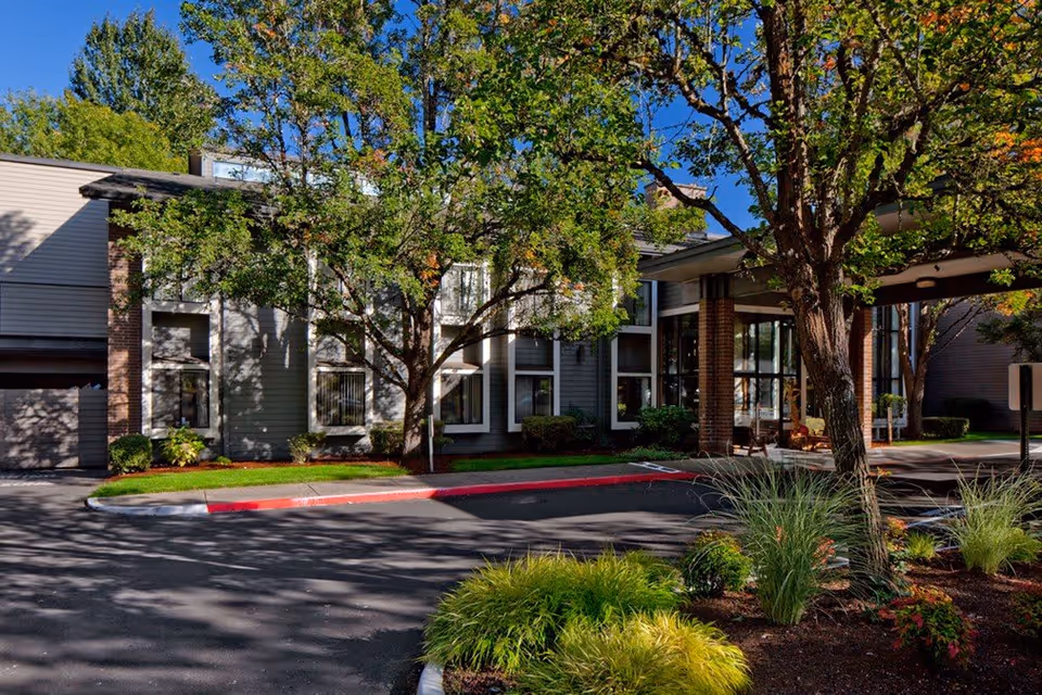 Exterior view of Creekside Village Retirement Residence showing a two-story building with large windows, surrounded by trees and landscaped greenery under a clear blue sky.