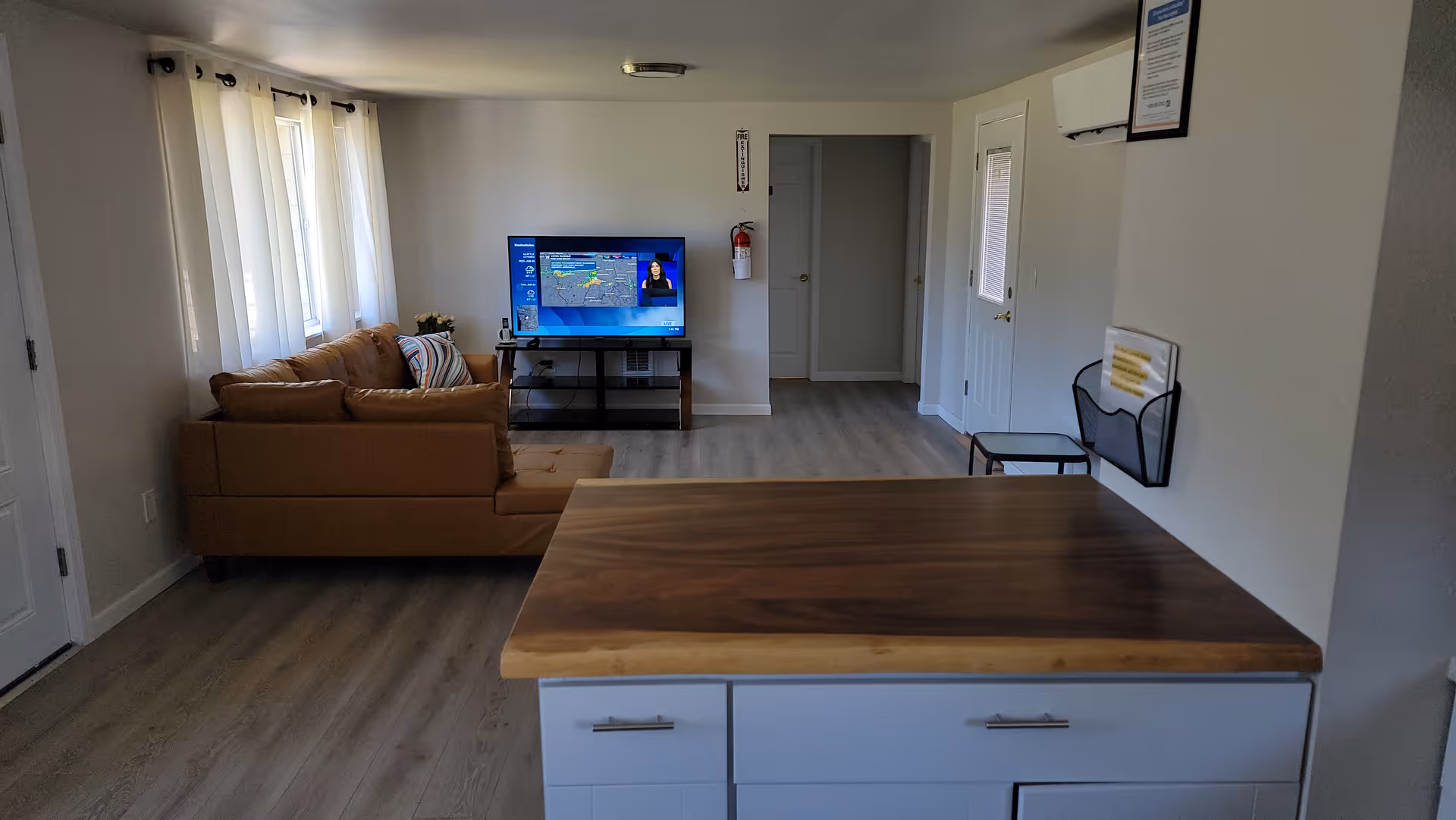 Interior view of a living room area with a brown sectional sofa facing a flat-screen TV on a stand. The room has light-colored walls and wood flooring. There are several windows with white curtains on the left side, and a wooden countertop with white cabinets in the foreground. A fire extinguisher is mounted on the wall near a doorway in the background.