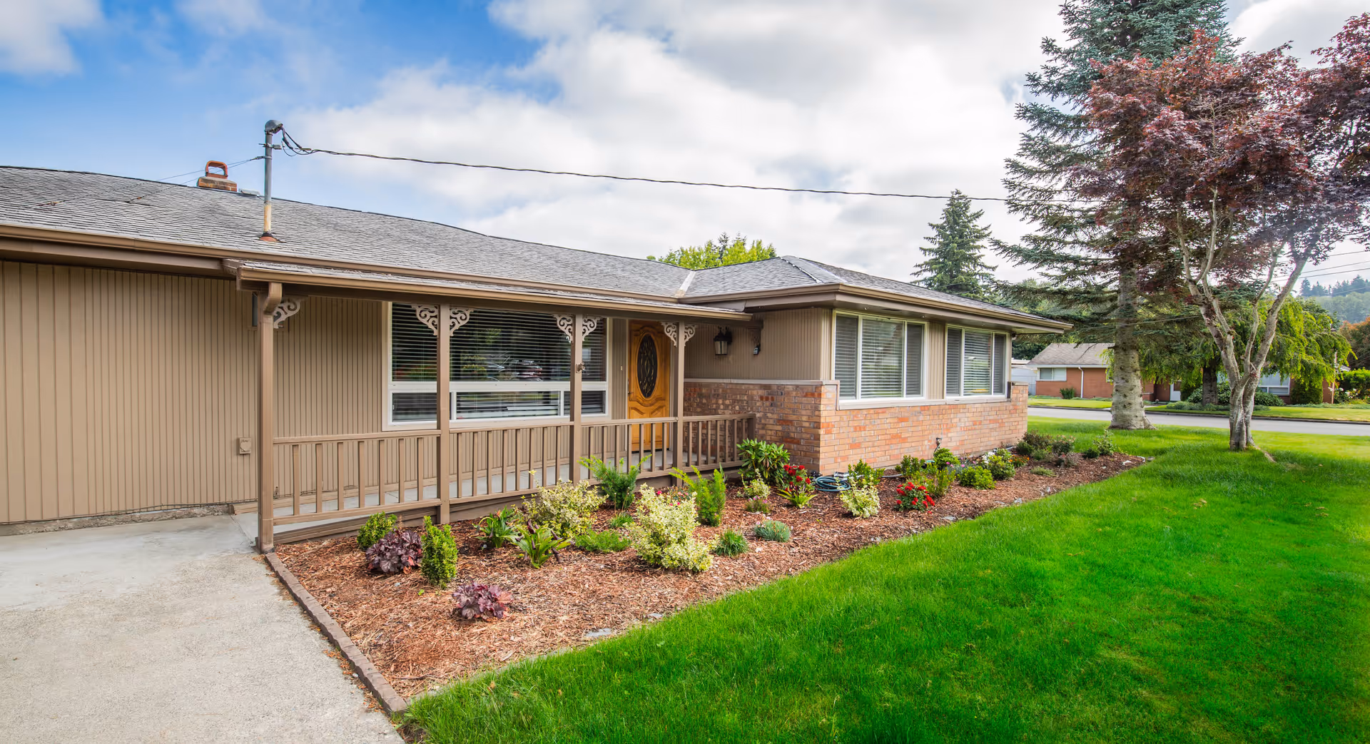 Exterior view of a single-story residential building with a brown wooden porch, a wooden front door with an oval glass panel, and large windows. The building has a combination of vertical siding and brickwork. In front of the porch is a landscaped garden bed with various shrubs and flowers, bordered by a green lawn. Trees and other houses are visible in the background under a partly cloudy sky.