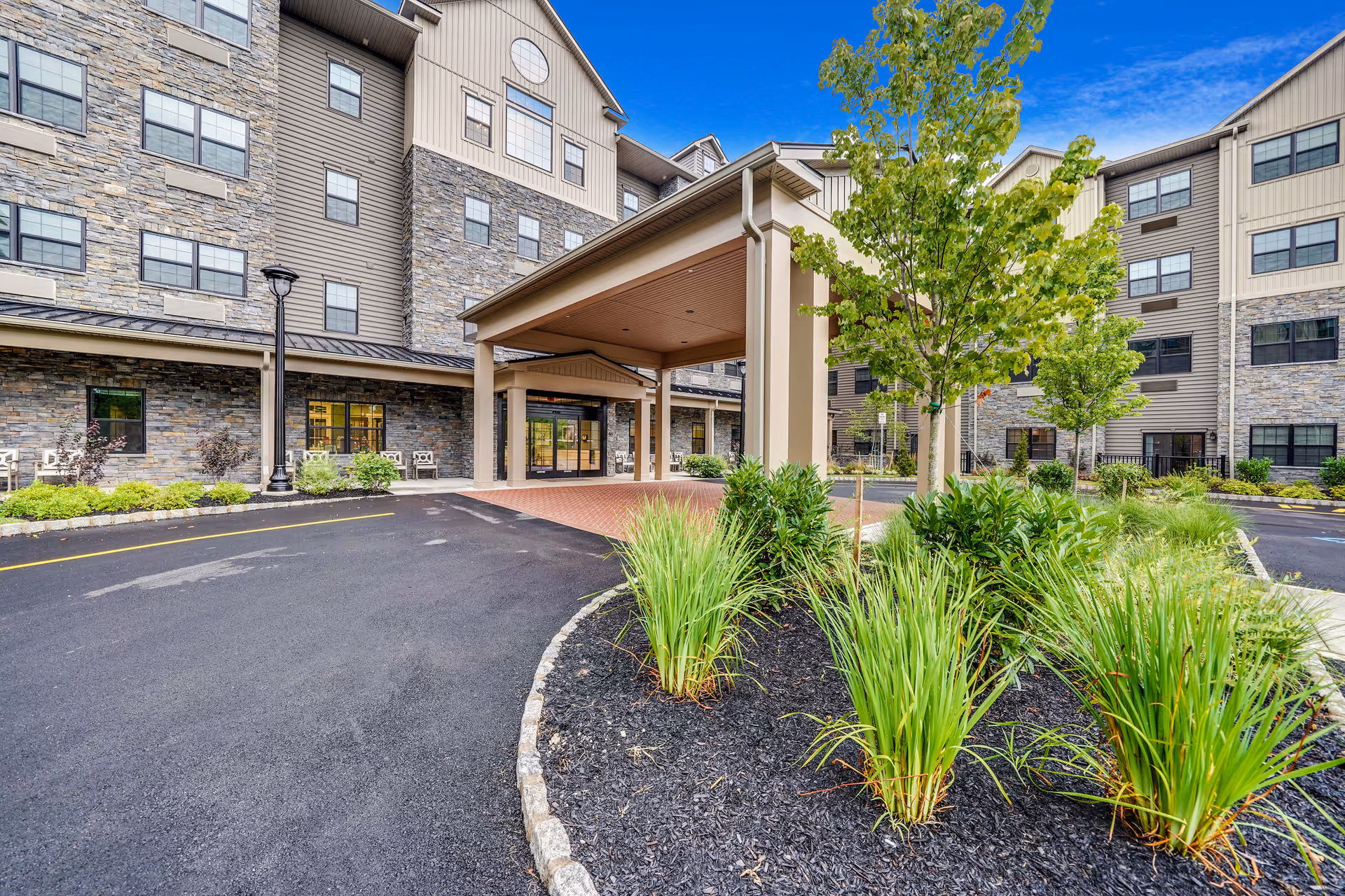Front entrance of a multi-story senior living building with a covered porte-cochere, driveway, and landscaped beds.