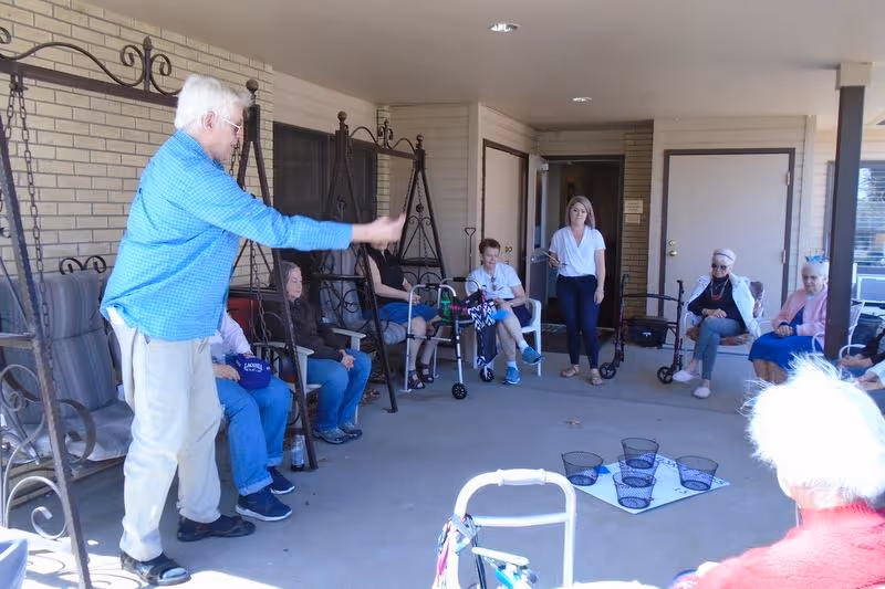 A group of seniors seated on a covered outdoor patio at an assisted living facility play a tossing game while a staff member watches.