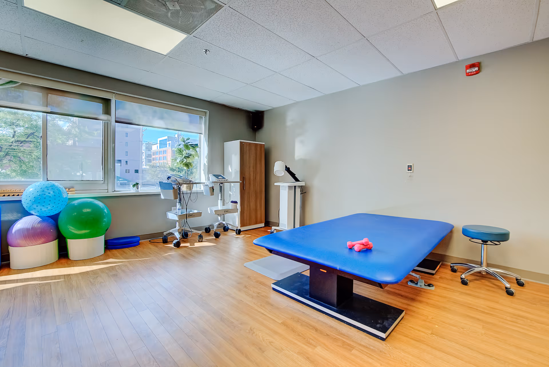 A physical therapy room with a blue therapy table, small pink dumbbells on the table, a rolling stool, exercise balls in various colors near the window, and medical equipment on carts. The room has large windows letting in natural light and wood flooring.