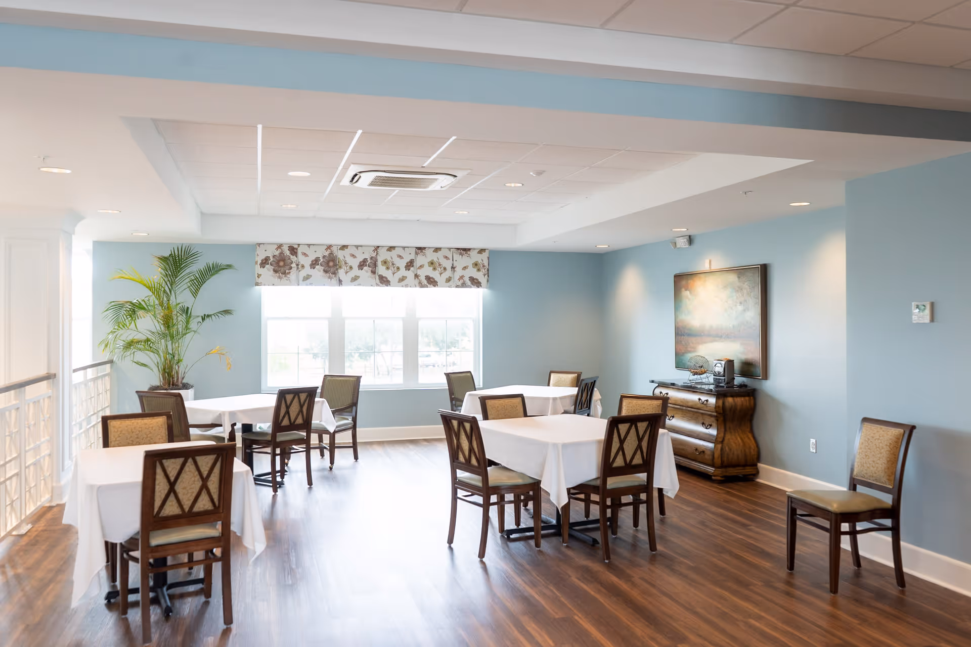 A bright dining room with light blue walls and wooden flooring. There are several tables covered with white tablecloths, each surrounded by wooden chairs with cushioned seats. A large window with floral valance lets in natural light. A potted plant is placed near the window, and a wooden sideboard with a decorative painting above it is against one wall.