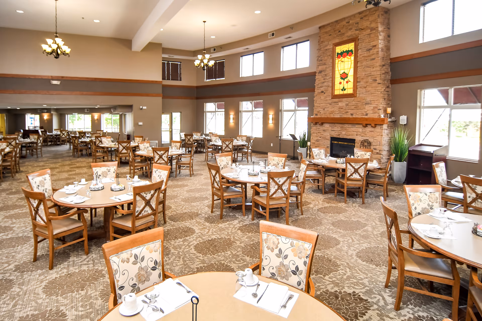 Spacious senior living dining room with multiple round tables and chairs, place settings, and a stone fireplace under tall windows.