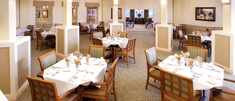A spacious dining room with multiple tables covered in white tablecloths, each set with plates, cups, napkins, and silverware. The room features beige walls, carpeted floors, and wooden chairs with cushioned seats and backs. There are decorative columns and framed artwork on the walls, with a seating area visible in the background.