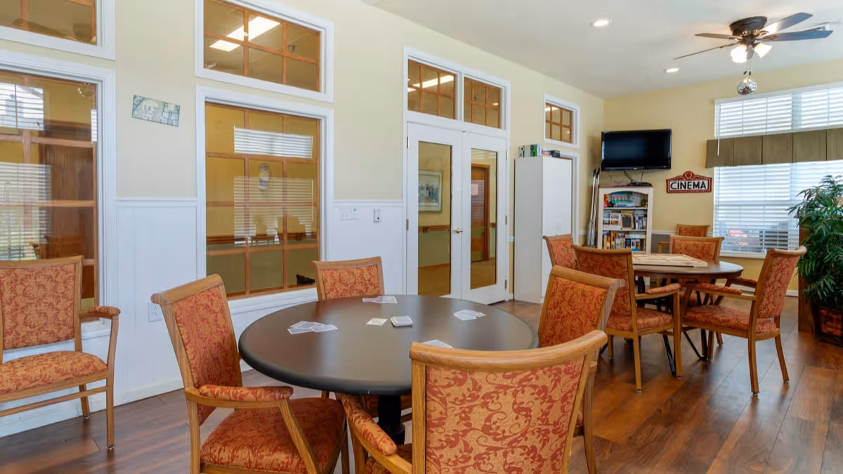 Bright common activity room with round tables and upholstered chairs, a TV and shelving near windows.