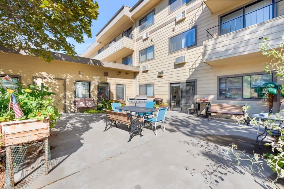 Outdoor courtyard with patio tables, chairs, planters and benches in front of a multi-story building exterior.
