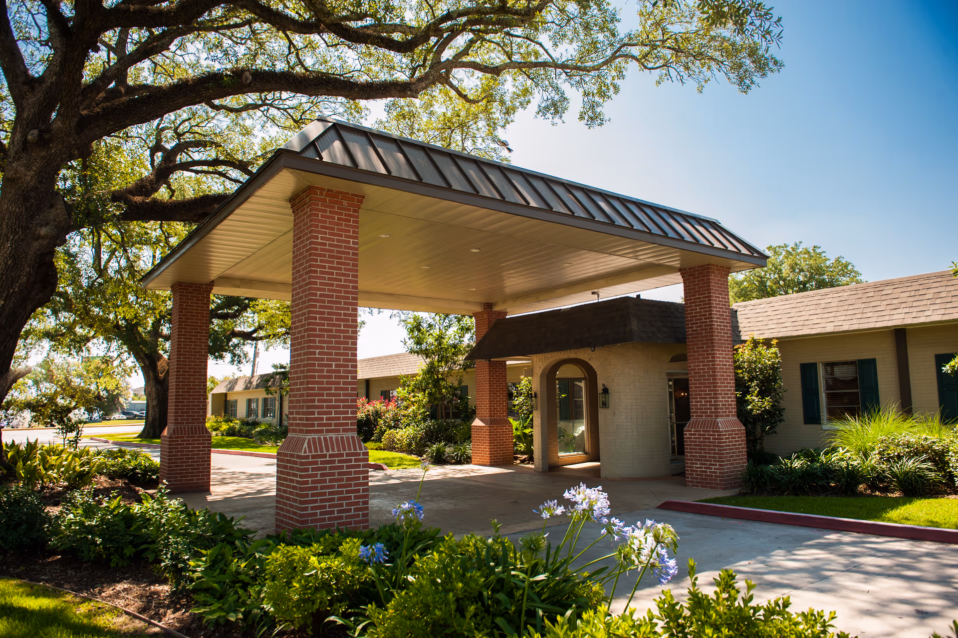 Entrance of Lady of the Oaks Retirement facility featuring a covered driveway supported by brick pillars, surrounded by well-maintained greenery and large trees under a clear blue sky.