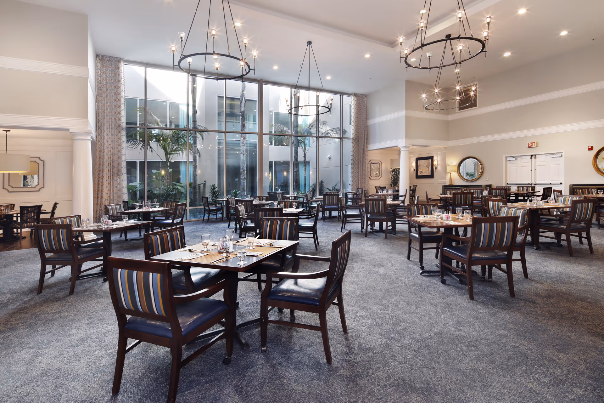 Spacious dining room with multiple set tables and striped chairs, large floor-to-ceiling windows, and hanging chandeliers.