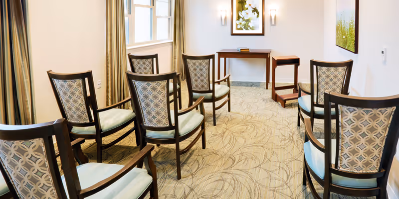 A small meeting or activity room with eight wooden chairs arranged in two rows facing a small wooden table against the wall. The room has patterned carpet, light-colored walls, two framed floral paintings, and windows with beige curtains allowing natural light.