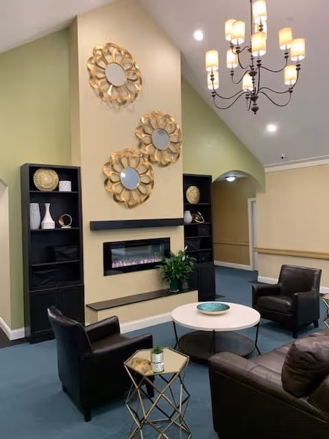 A cozy living room area with a modern electric fireplace set into a beige wall. Above the fireplace are three decorative round mirrors with gold petal-like frames. On either side of the fireplace are black shelving units with decorative items. The room has a high ceiling with a chandelier featuring multiple lights. There are three dark leather armchairs and a matching sofa arranged around a white round coffee table and a small gold side table with a plant on it. The walls are painted in soft green and beige tones.