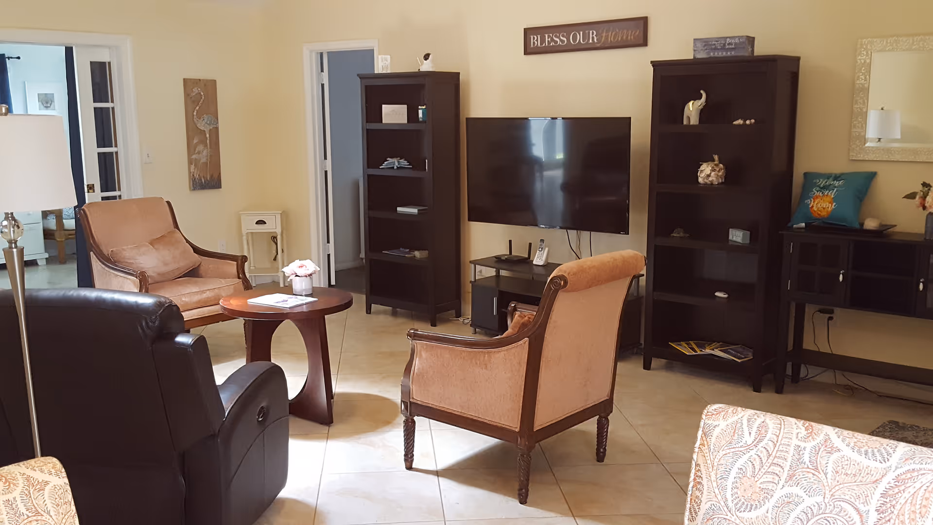 Cozy living room with upholstered chairs around a small table, a wall-mounted TV flanked by dark bookshelves, and a decorative 'Bless Our Home' sign above.
