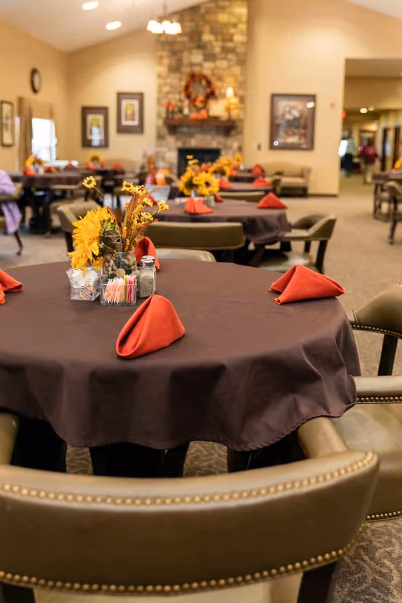 Dining area in a senior living facility with round tables covered in dark tablecloths, each set with red folded napkins and a centerpiece featuring sunflowers and other decorative elements. In the background, there is a stone fireplace decorated with a wreath and framed pictures on the walls.