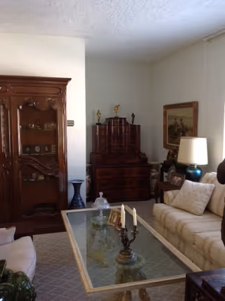 A cozy living room with a beige sofa adorned with pillows, a glass coffee table with decorative items including a candelabra and framed photos, a wooden cabinet with shelves displaying various items, a dark wooden chest of drawers with small statues on top, a table lamp with a white shade, and a framed painting on the wall.