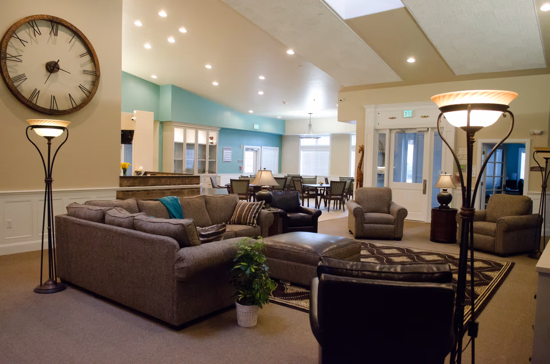 A spacious and well-lit living room area in an assisted living facility featuring a large gray sectional sofa, two armchairs, a black leather chair, a large ottoman, two tall floor lamps, a large wall clock, and a patterned rug. In the background, there is a dining area with tables and chairs, a reception desk, and large windows with blinds.