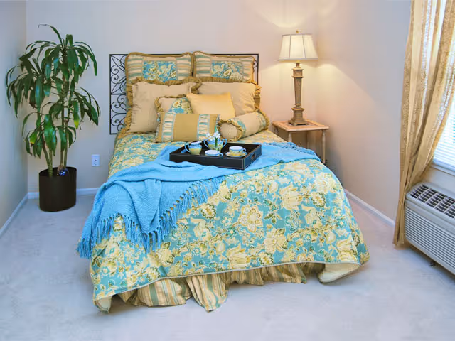 A cozy bedroom featuring a bed with blue and yellow floral bedding, a bedside lamp, a potted plant, and a window air conditioner.