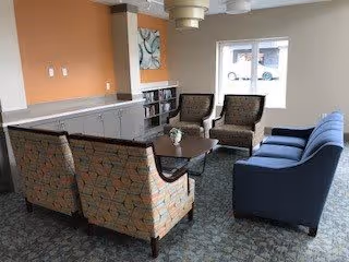 A cozy seating area in a senior living facility with two patterned armchairs, two solid-colored armchairs, and a dark wooden coffee table in the center. The room has a window letting in natural light, a built-in cabinet with shelves, and a piece of abstract wall art above the cabinet.