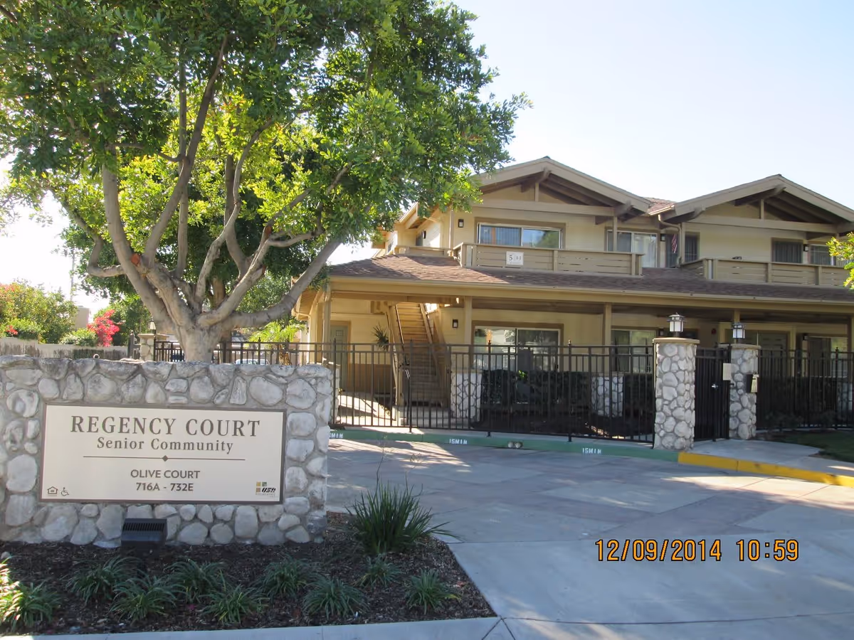 Exterior view of Regency Court Senior Community showing a two-story building with balconies, a gated entrance, and a stone sign in front that reads 'REGENCY COURT Senior Community OLIVE COURT 716A - 732E'. There are trees and landscaping around the sign and building.