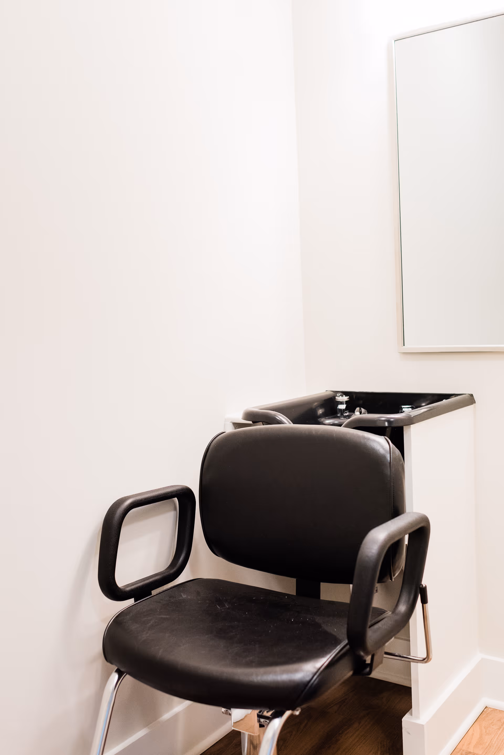 A black salon chair with armrests positioned in front of a black sink basin in a small, clean room with white walls and a mirror on the wall.