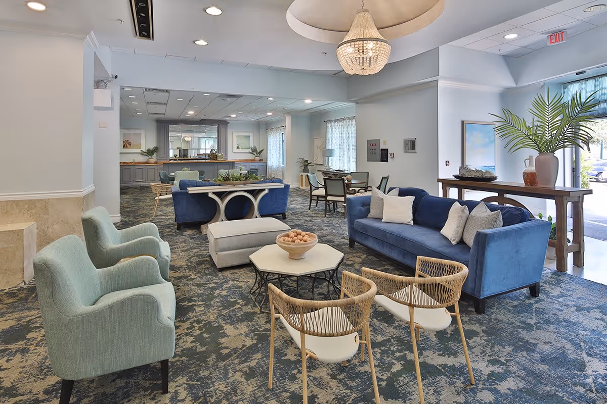 A spacious and well-lit living room area in The Sterling Aventura facility featuring blue sofas, light green armchairs, two wicker chairs, a hexagonal coffee table with a decorative bowl, and a chandelier hanging from the ceiling. There is a console table with a large plant near the entrance and a dining area with chairs and tables in the background.