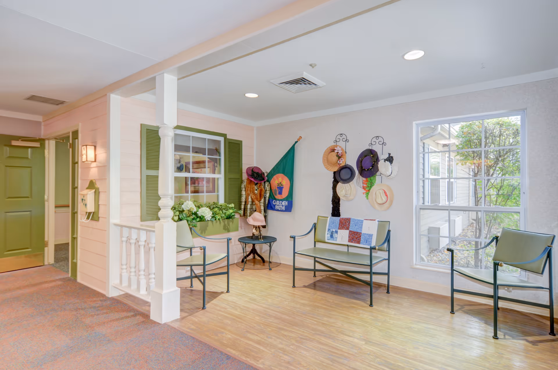 A cozy indoor seating area with three green metal chairs and a matching bench. The bench has a colorful quilt draped over it. On the wall above the bench, there are decorative hat racks holding various hats and scarves. A green flag with the words 'Garden Path' is hung on the wall. There is a window with green shutters and a flower box filled with white flowers. The floor is a combination of wood and carpet, and a large window lets in natural light, showing some greenery outside.