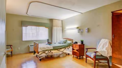 A hospital-style bedroom with adjustable bed, nightstand, chair, and window in a senior facility.