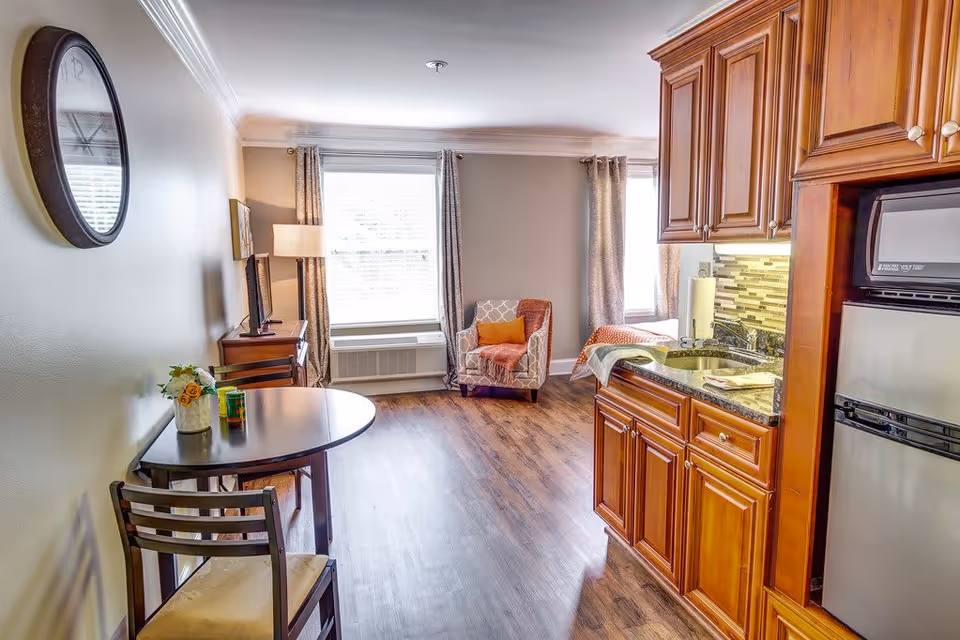 A bright and cozy living space featuring a small kitchen area with wooden cabinets, a granite countertop, a sink, a microwave, and a mini refrigerator. Adjacent to the kitchen is a round dining table with two chairs, a vase with flowers, and a can on the table. The room has wooden flooring, two large windows with curtains, a comfortable armchair with a throw blanket and pillow, a TV on a wooden stand, a floor lamp, and a wall clock.
