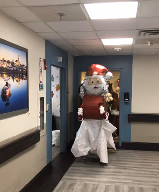 A hallway in a senior living facility decorated with a large Santa Claus figure wearing a red hat and holding a red box. The hallway has light-colored walls with a blue accent wall at the end, ceiling lights, and a framed picture on the left wall. The floor has a wood-like pattern.