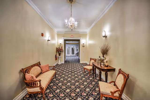A long hallway with patterned carpet and beige walls, furnished with vintage wooden chairs and a small table with decorative items. A chandelier hangs from the ceiling, and there are wall sconces providing light. At the end of the hallway, double doors are visible.