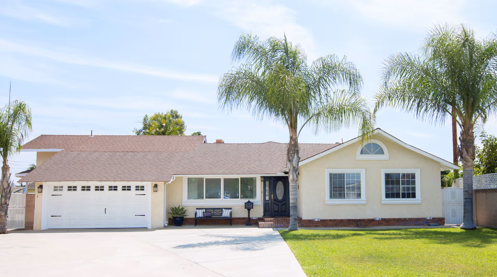 Single-story beige house with a brown shingle roof, a white garage door, a black front door, two palm trees in the front yard, a green lawn, and a bench near the window under a clear blue sky.