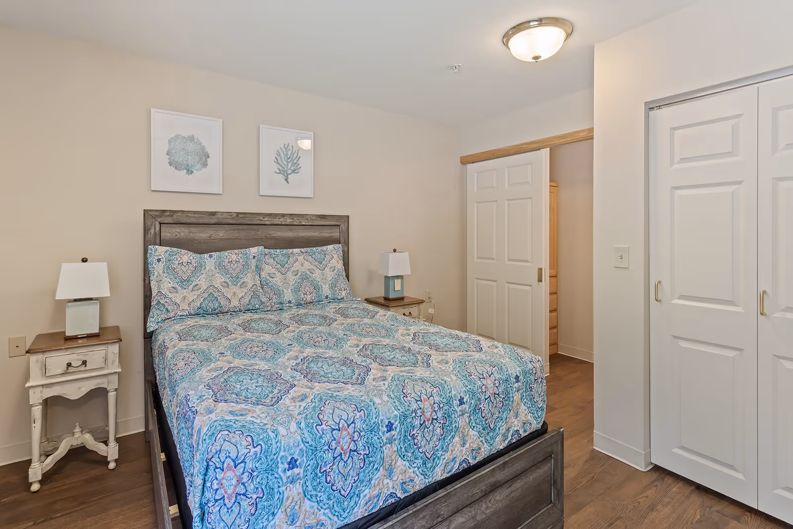 Well-lit bedroom with a patterned blue bedspread, wooden headboard, two nightstands with lamps, and white closet doors.