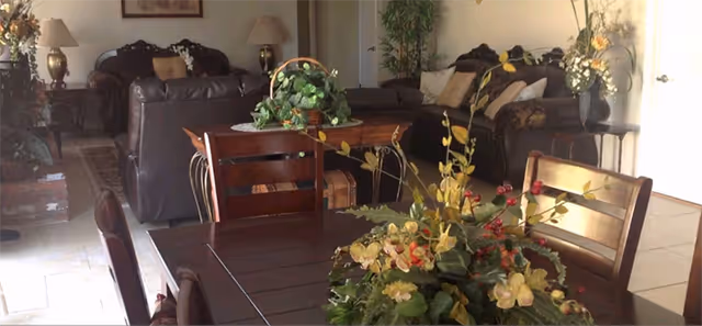 A cozy living room and dining area with wooden furniture, including a dining table with chairs and a floral centerpiece. The living room features dark leather sofas, decorative pillows, table lamps, and various plants and floral arrangements, creating a warm and inviting atmosphere.