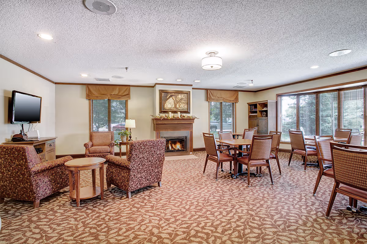 A spacious senior living common area with patterned carpet, several wooden tables and chairs, two upholstered armchairs around a small round wooden table, a fireplace with a painting above it, large windows with brown valances, a wall-mounted flat screen TV, and a wooden cabinet with a lamp and plant.