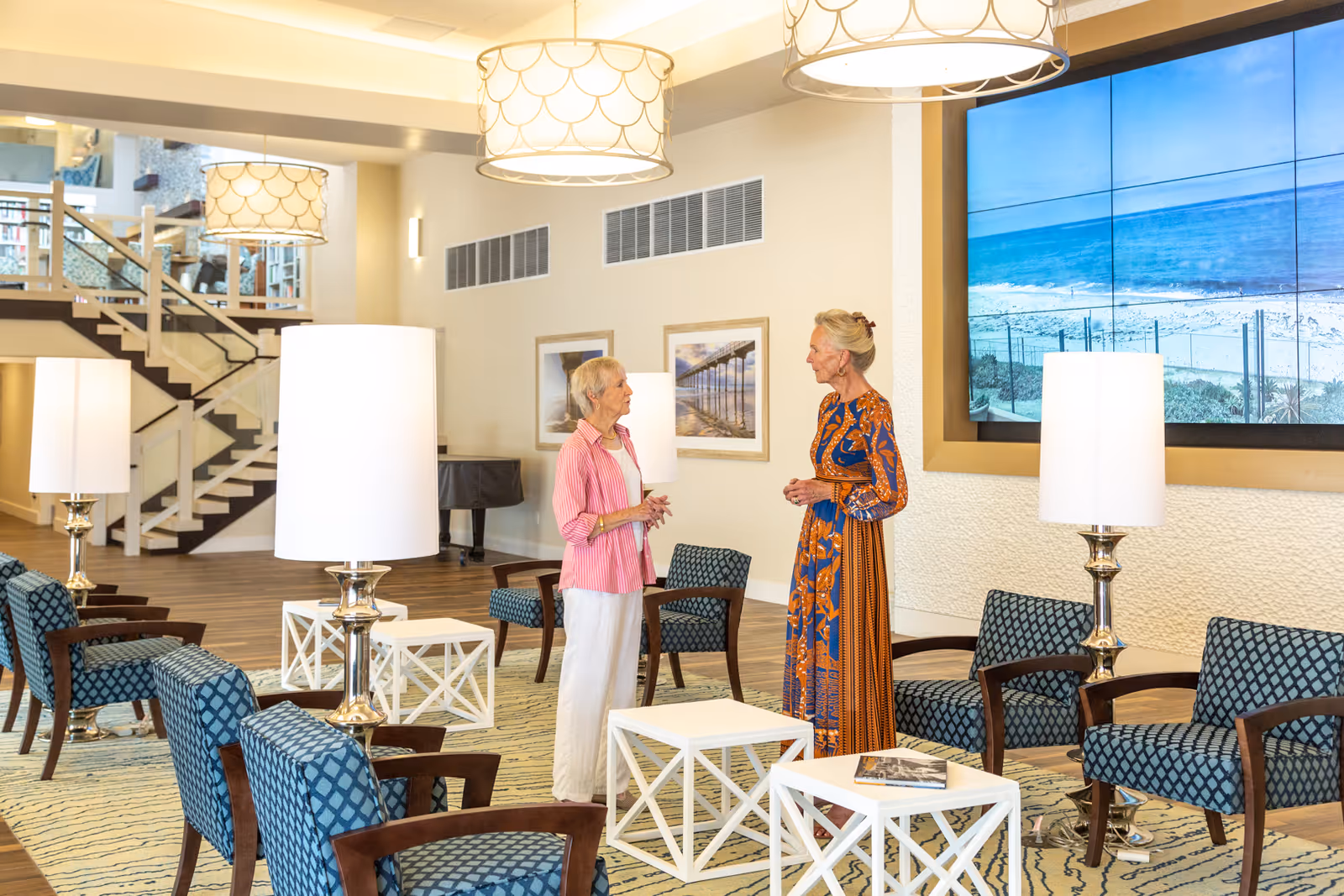 Two women talking in a bright senior living lounge with patterned chairs, white side tables, tall lamps, a large wall screen showing a beach, and a staircase in the background.