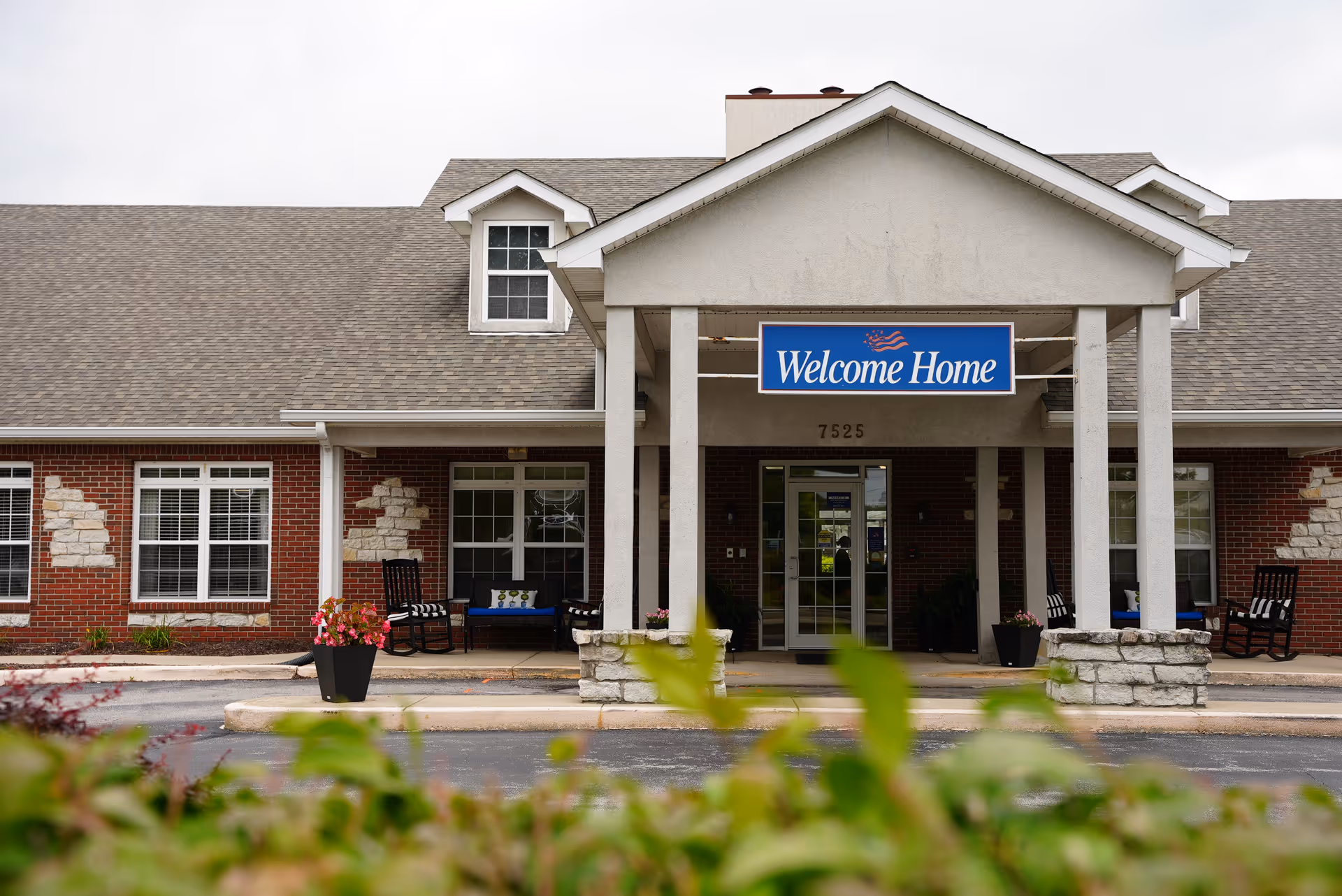 Exterior front entrance of a senior living facility with a covered porch, brick walls with stone accents, several windows, and a blue sign that reads 'Welcome Home' above the doorway. There are chairs and potted plants on the porch, and greenery is visible in the foreground.