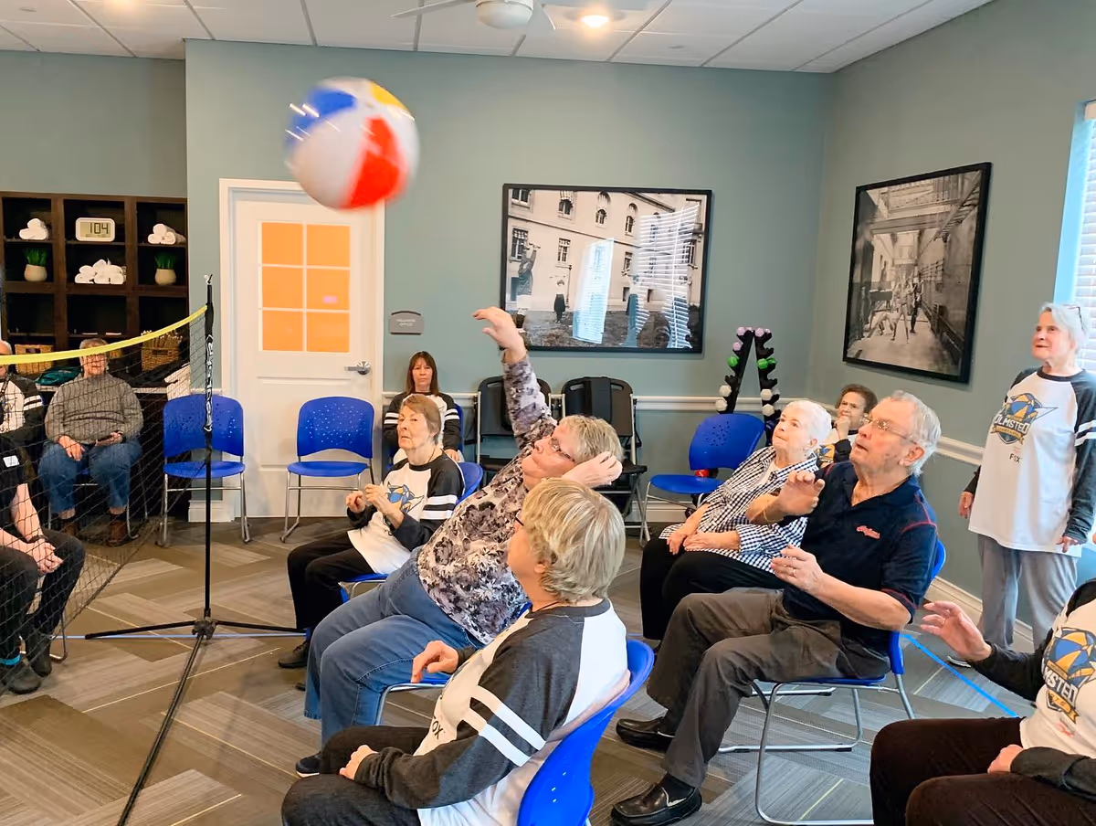 A group of elderly people seated in a room playing a seated volleyball game with a colorful beach ball over a low net. The room has blue chairs, framed black and white photos on the walls, and a rack of small dumbbells in the corner.