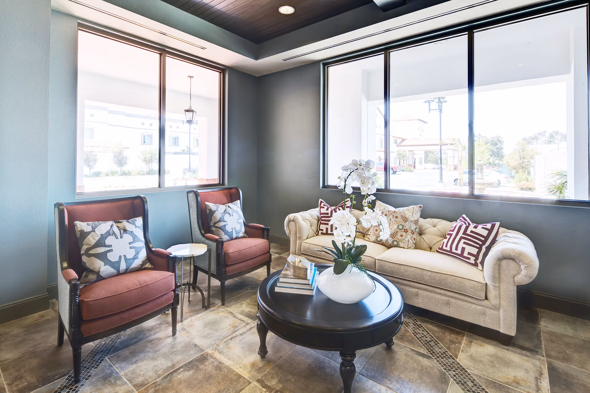 Sunlit lounge with a beige tufted sofa, two red armchairs, a round dark coffee table with a white vase of orchids, and large windows.