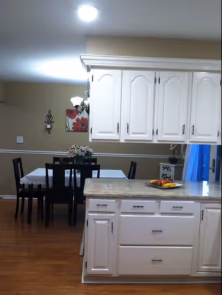 Interior view of a senior living facility showing a kitchen island with white cabinets and a granite countertop. In the background, there is a dining area with a table covered by a white tablecloth and six black chairs. A chandelier hangs above the dining table, and a floral wall decoration is visible on the beige wall. The floor is wooden.