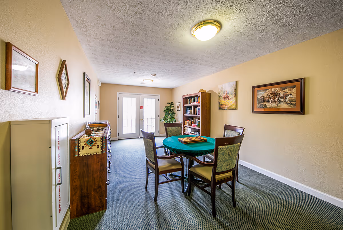 A well-lit room with a round table covered by a green tablecloth and four chairs around it. There is a bookshelf filled with books against the far wall, two framed paintings on the right wall, and a wooden cabinet with a decorative cloth on the left side. Double glass doors at the end of the room let in natural light.